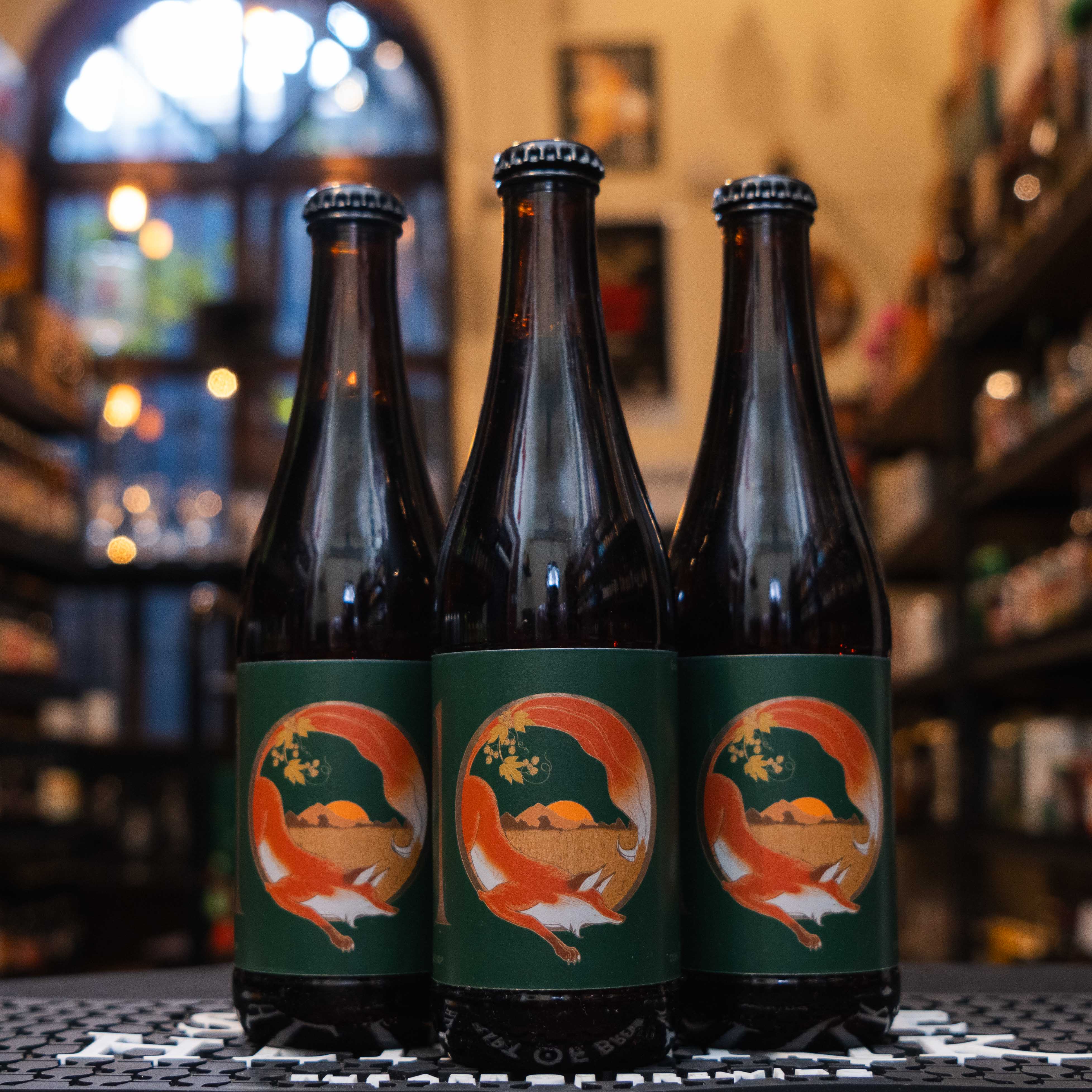 Botella de La Zorra 10 Aniversario, una Imperial IPA estilo Citra Single Hop. La etiqueta es verde con una ilustración central de una zorra roja rodeando un paisaje con lúpulos dorados. Con un ABV de 8.5%, ofrece intensas notas cítricas y tropicales a toronja, mango y piña, con un amargor robusto. Ideal para maridar con comida picante y quesos añejos. Su diseño celebra una década de excelencia cervecera.