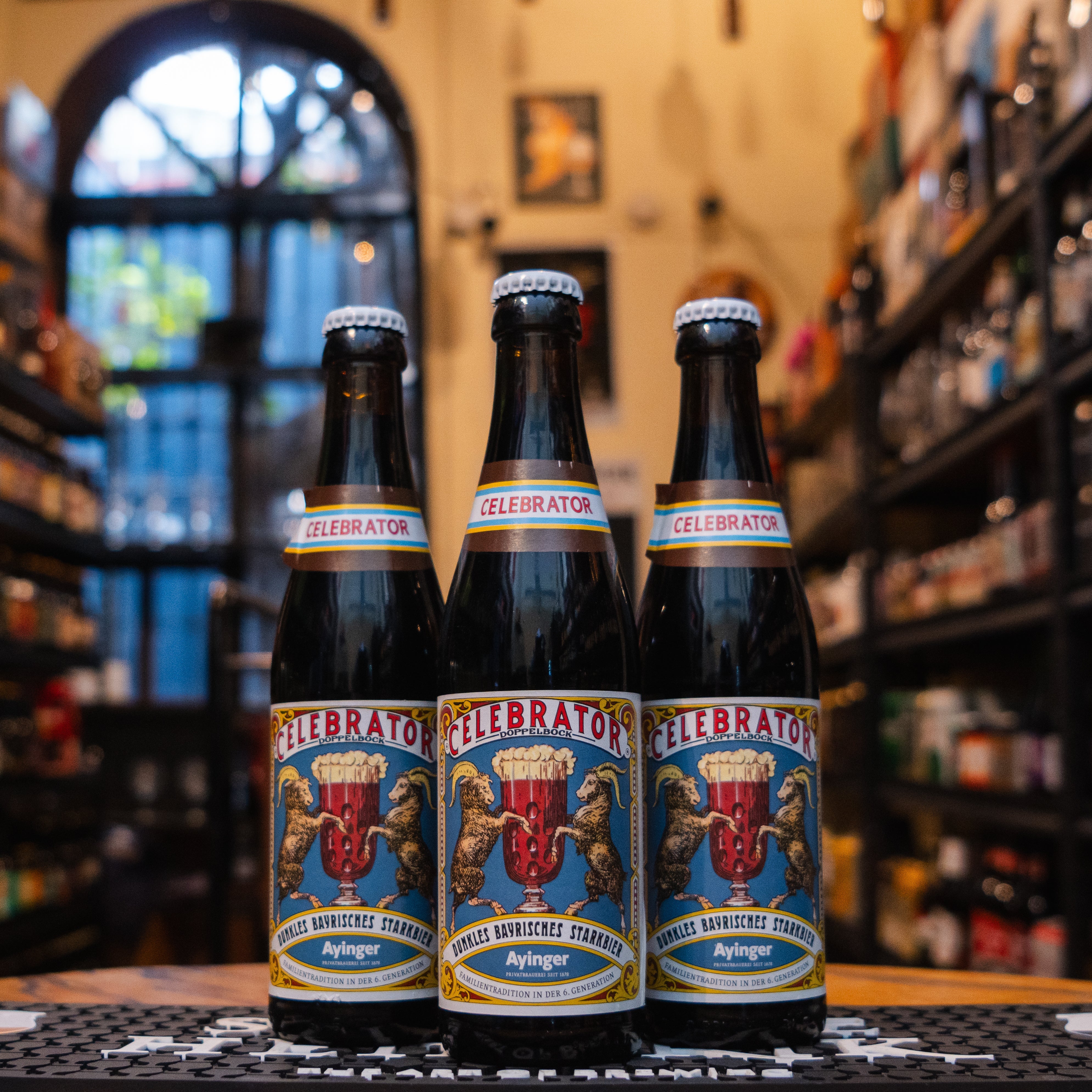 Botella de Ayinger Celebrator Doppelbock, una cerveza bávara tipo Doppelbock con 6.7% ABV. La etiqueta muestra un fondo azul, dos cabras rodeando una copa y detalles dorados. El nombre 'Celebrator' destaca en banda roja. Sabor a caramelo, frutos secos y malta tostada. Ideal con carnes asadas, guisos o strudel de manzana. Diseño clásico que refleja tradición cervecera alemana.