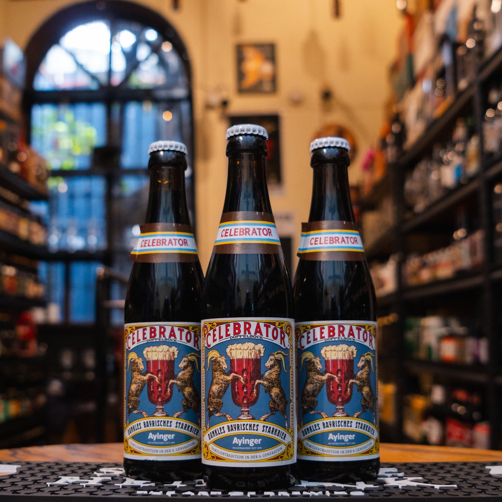 Botella de Ayinger Celebrator Doppelbock, una cerveza bávara tipo Doppelbock con 6.7% ABV. La etiqueta muestra un fondo azul, dos cabras rodeando una copa y detalles dorados. El nombre 'Celebrator' destaca en banda roja. Sabor a caramelo, frutos secos y malta tostada. Ideal con carnes asadas, guisos o strudel de manzana. Diseño clásico que refleja tradición cervecera alemana.