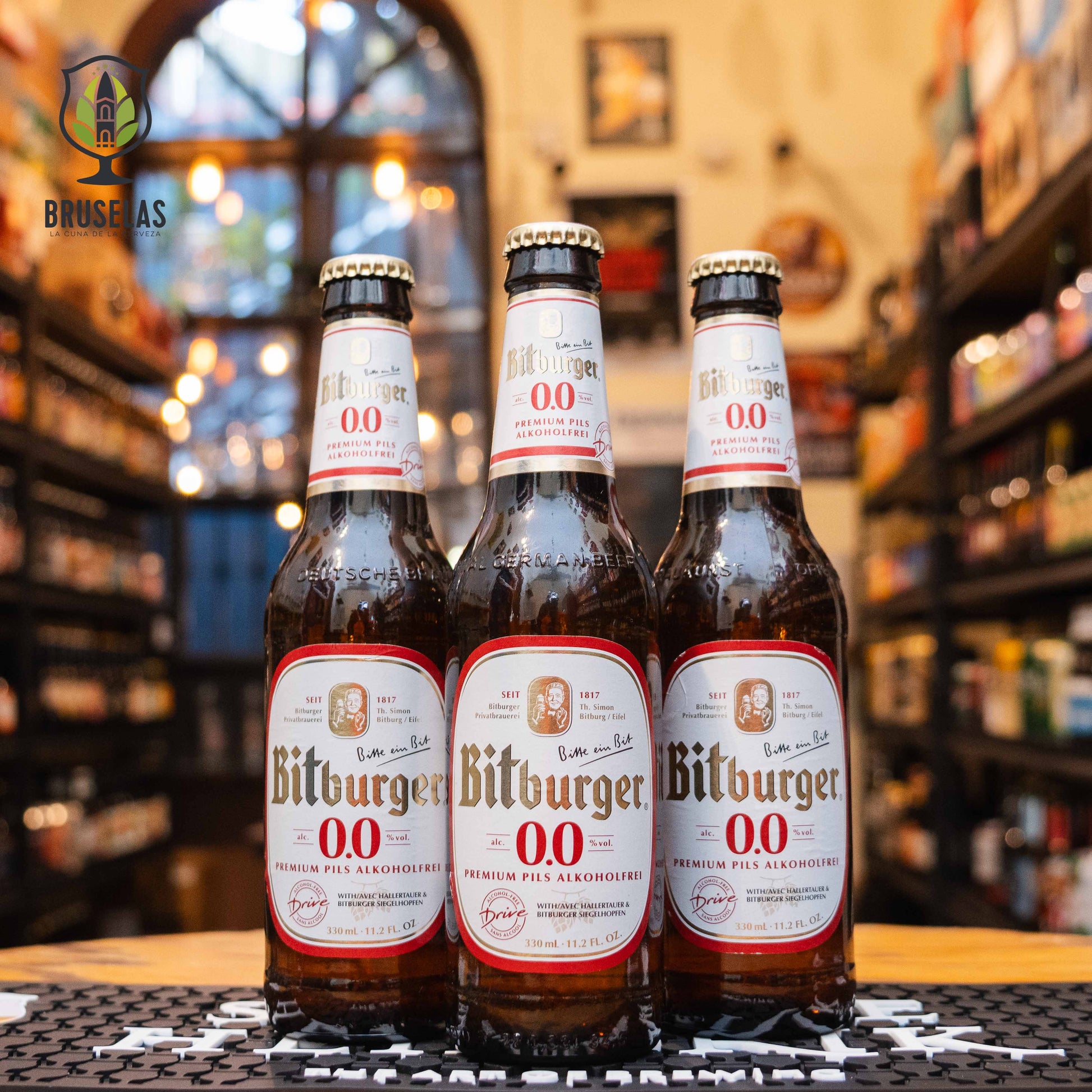 Botella de Bitburger Drive 0.0%, una pilsner sin alcohol de Bitburger Braugruppe. La etiqueta es blanca con detalles rojos y dorados, con el nombre de la marca en letras doradas. ABV de 0.0%, con un sabor refrescante, equilibrado entre dulzura malteada y amargor suave. Ideal para acompañar ensaladas y platos ligeros. El diseño de la botella de 330 ml refleja la pureza de esta cerveza sin alcohol.