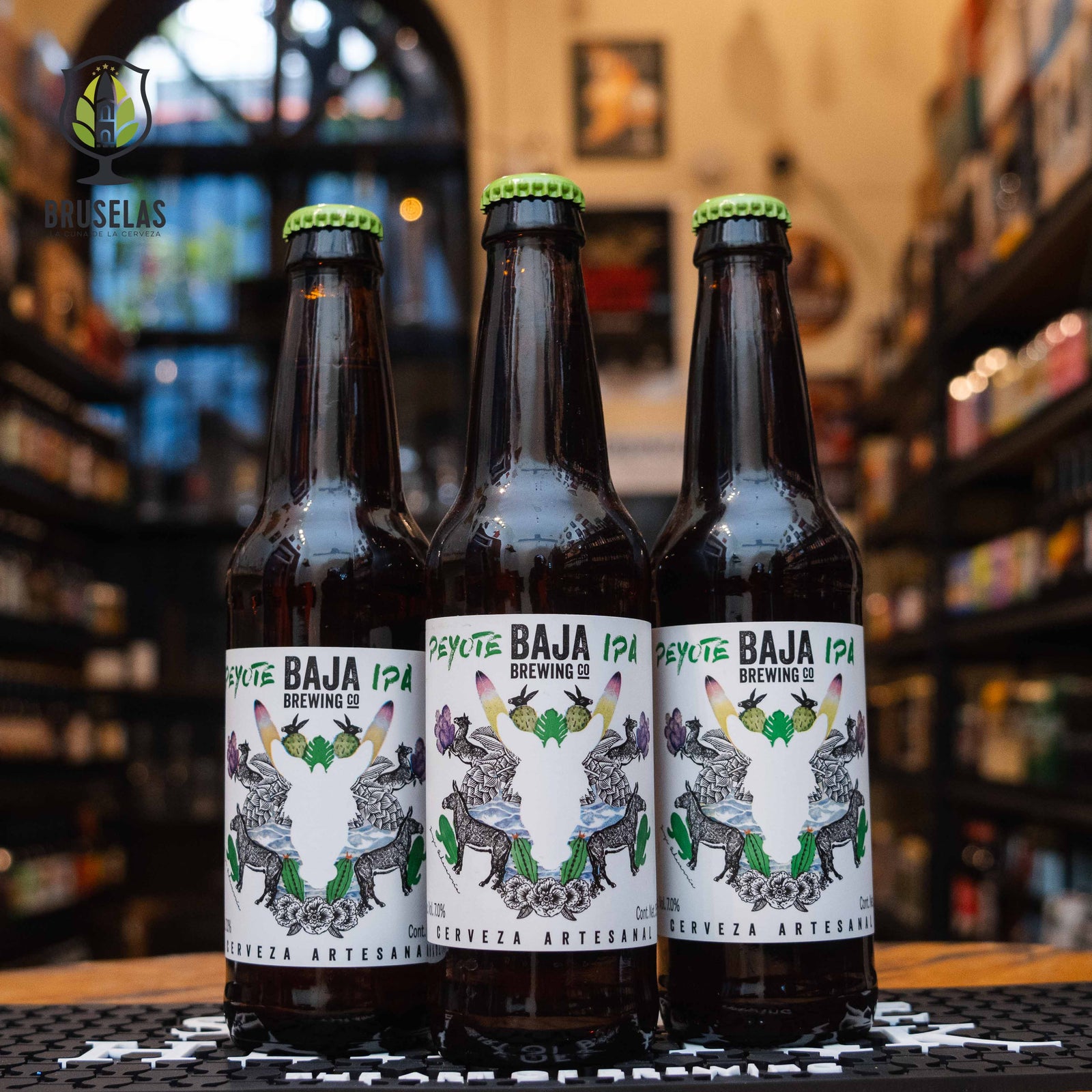 Botella de Baja Peyote IPA, una American IPA de Baja Brewing Co. La etiqueta tiene un diseño de venado con cactus y flores, con colores predominantes verde, blanco, naranja y púrpura. El nombre 
