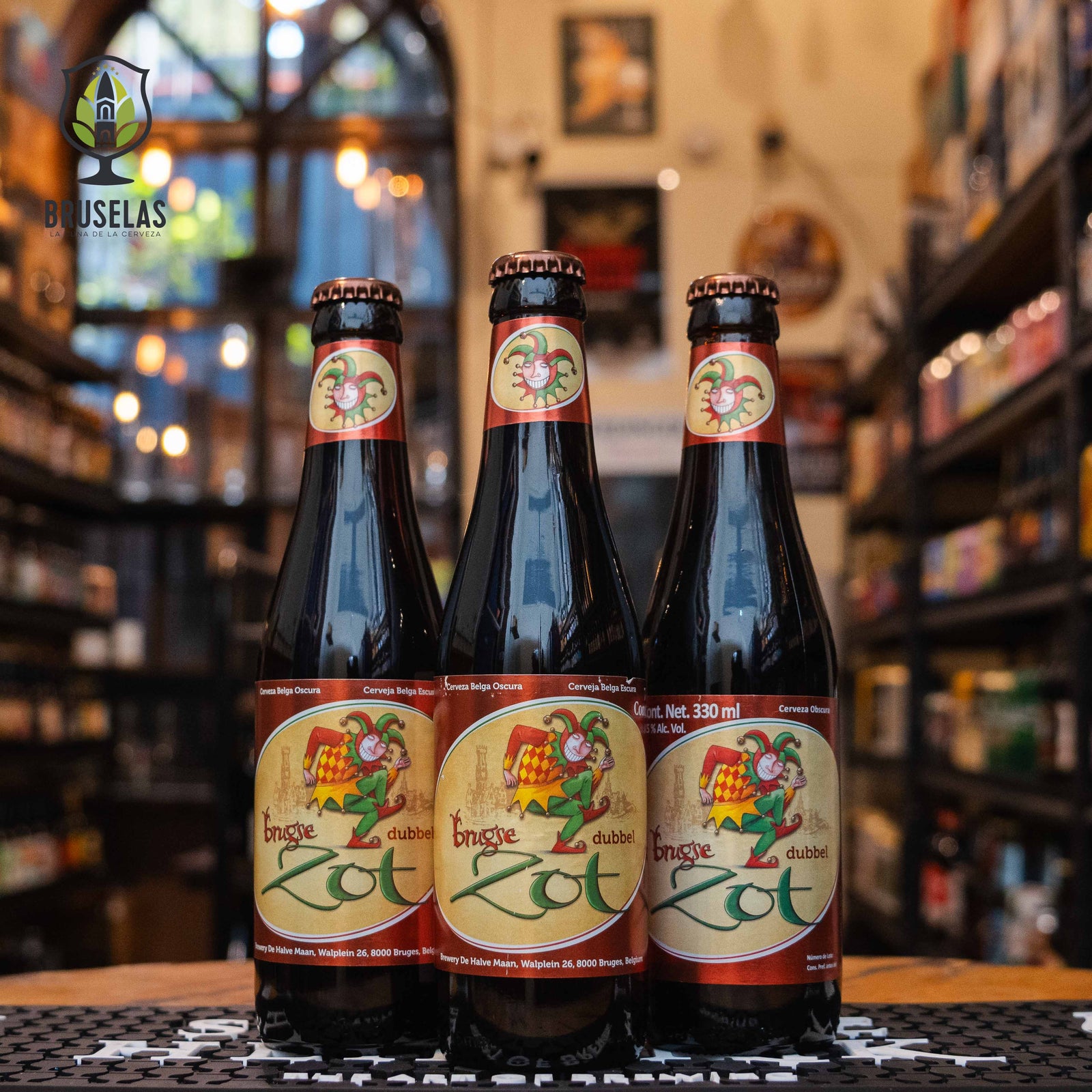 Botella de Brugse Zot Dubbel, una Belgian Dubbel elaborada por Brouwerij De Halve Maan en Brujas, Bélgica. La etiqueta muestra un diseño clásico con un bufón, reflejando su historia. Con un ABV de 7.5%, ofrece aromas de miel, almendras, chocolate y azúcar moreno. Su sabor es complejo, con malta tostada, caramelo, pasas y ciruelas, y un final ligeramente amargo. Ideal para acompañar estofados de carne, quesos variados y postres de chocolate amargo. El diseño evoca la rica tradición belga.