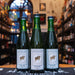 Botella de Cantillon Gueuze, una cerveza belga tipo gueuze con 5% ABV, elaborada por Brasserie Cantillon. La etiqueta es blanca con un diseño minimalista, destacando un cordero dorado en el centro y líneas radiantes alrededor. Ofrece un perfil ácido con notas frutales y amaderadas. Ideal para maridar con quesos curados, pescados ahumados y postres cítricos. Su diseño refleja tradición artesanal belga.