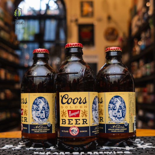 Botellas de Coors Banquet en una tienda de cerveza artesanal. Esta American Lager, elaborada con agua de las Montañas Rocosas, ofrece un sabor refrescante y es perfecta para maridar con hamburguesas, pizzas y snacks salados. Su icónica botella y presencia en series como Yellowstone la convierten en una cerveza de culto. ¡Disfrútala bien fría!