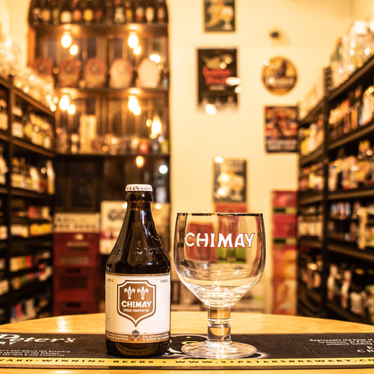 Copa Chimay 330ml, de cristal en forma de cáliz con logo rojo y blanco. Ubicada sobre una barra junto a una botella de Chimay Triple, resaltando su uso ideal para cervezas trapenses.