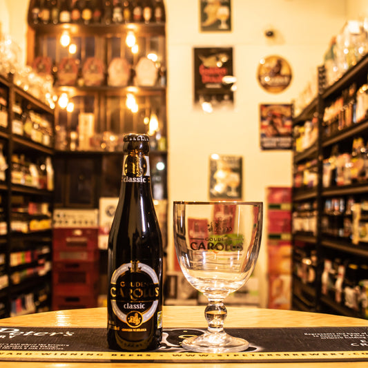  Copa Gouden Carolus 330ml, de cristal en forma de cáliz con logo y borde dorado. Ubicada sobre una barra junto a una botella de Gouden Carolus Classic, destacando su uso ideal para cervezas belgas.