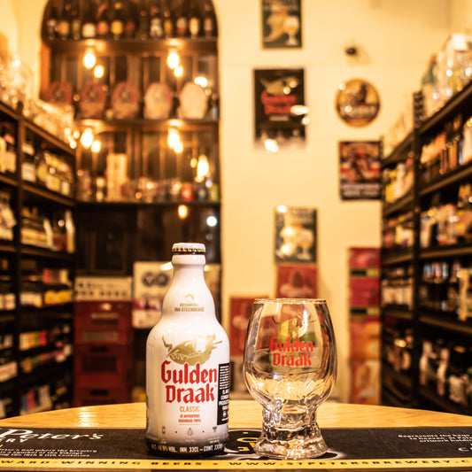 Copa Gulden Draak 500ml, con diseño abombado y base sólida, decorada con el logo rojo y dorado. Ubicada sobre una barra junto a una botella de Gulden Draak Classic, resaltando su uso ideal para degustaciones cerveceras exclusivas.