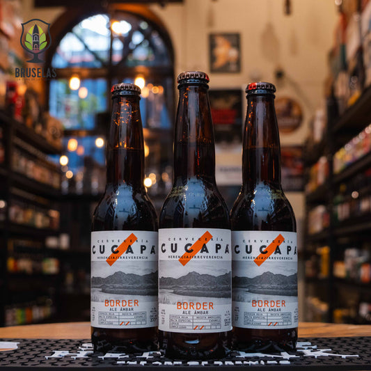 Botella de Cucapá Border, una Amber Ale de Cervecería Cucapá, elaborada en Mexicali, Baja California. La etiqueta tiene un diseño en tonos gris y blanco, con una imagen de montañas y un logotipo naranja en diagonal. El nombre aparece en letras negras y naranjas. Con un ABV de 4.5%, ofrece notas de caramelo y miel con un amargor ligero. Ideal para acompañar carnes asadas o tacos al pastor. El diseño refleja una estética moderna y minimalista.