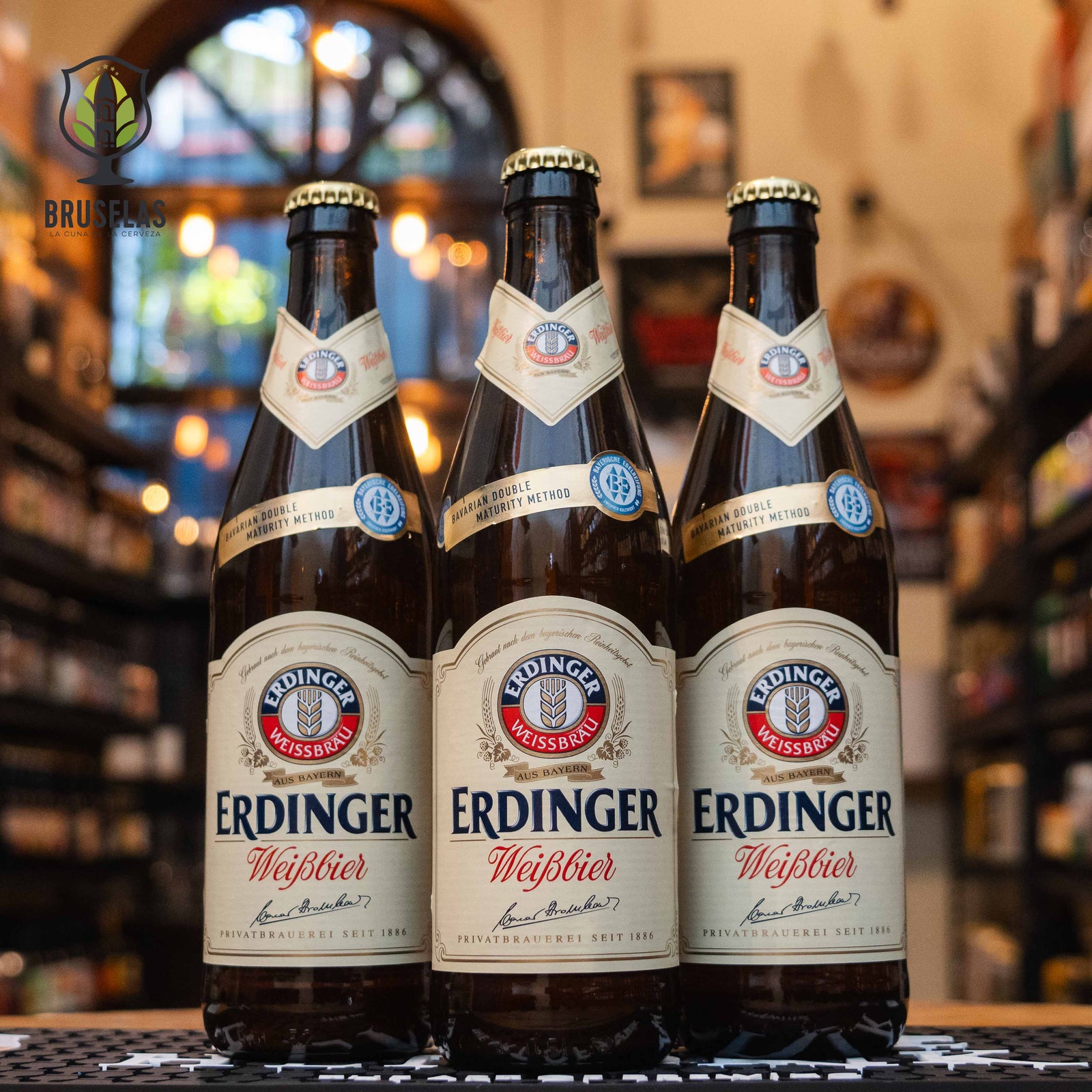 Botella de Erdinger Weissbier, una reconocida cerveza de trigo elaborada por Erdinger Weissbräu en Baviera, Alemania. Con un ABV de 5.3%, esta Weissbier se caracteriza por su color dorado turbio y espuma densa. Ofrece aromas a plátano, clavo y vainilla, mientras que su sabor es suave, frutal y ligeramente especiado, con un final refrescante. Ideal para maridar con platos de pescado, ensaladas ligeras y quesos suaves. Presentación en botella de 500 ml.