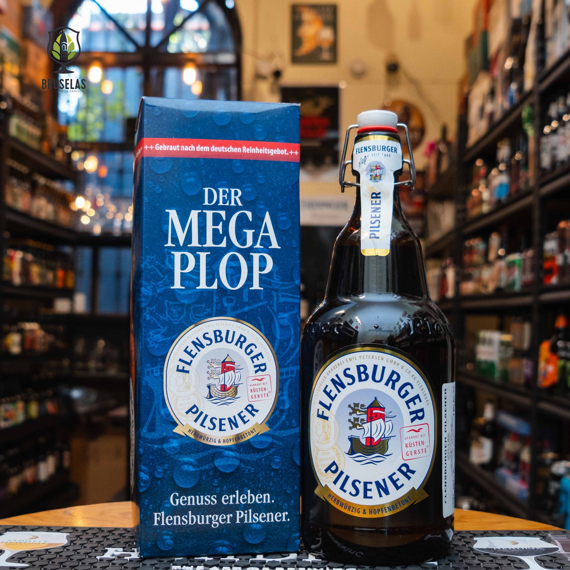 Botella Flensburger Pilsener Mega Plop de 2L, una Pilsner alemana de 4.8% ABV, junto a su empaque azul con el logo de la cervecería. Ideal para compartir y maridar con mariscos, pretzels y embutidos.