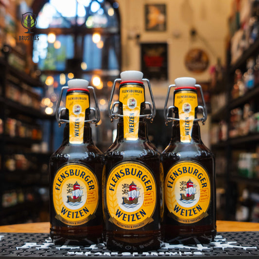 Botella de Flensburger Weizen, una Hefeweizen alemana de 5.1% ABV. Su etiqueta amarilla con el icónico barco y su tapón mecánico resaltan su tradición cervecera. Ideal para acompañar salchichas alemanas, pretzels y quesos suaves.