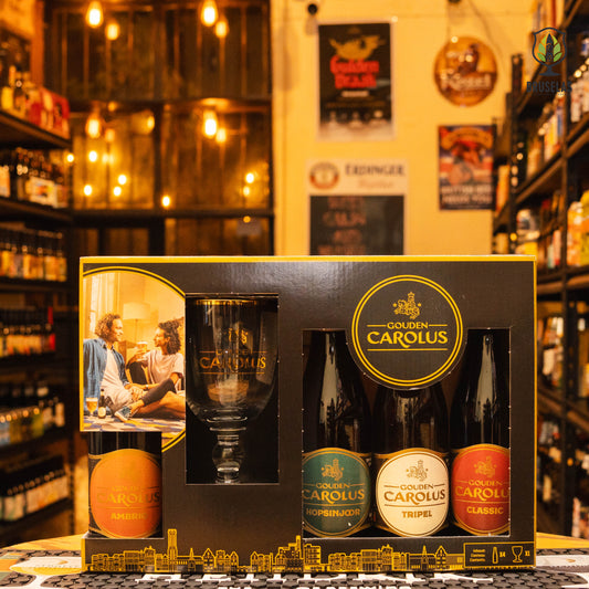 Giftpack Gouden Carolus se presenta en una elegante caja negra con detalles dorados, destacando la marca y su herencia belga. Se pueden ver cuatro botellas de distintos estilos y una copa oficial con el logo Gouden Carolus, diseñada para una experiencia de degustación óptima. El fondo muestra un ambiente acogedor con iluminación cálida y estanterías llenas de cervezas artesanales. Cambios sujetos a disponibilidad.