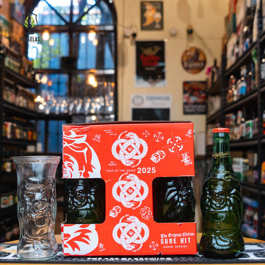 El Giftpack Lucky Buddha se presenta en una llamativa caja roja con ilustraciones del Buda de la fortuna y detalles inspirados en la cultura china. A través de la ventana de la caja se pueden ver las icónicas botellas de vidrio verde con forma de Buda. A un lado, un vaso exclusivo con el mismo diseño. El fondo muestra una tienda de cervezas artesanales con estanterías bien surtidas e iluminación cálida. Cambios sujetos a disponibilidad.