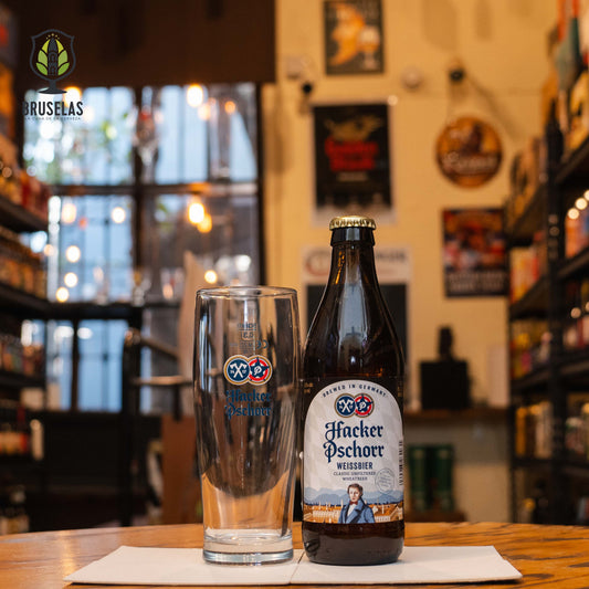 Vaso Hacker-Pschorr 300ml, de cristal alto y estilizado con logo azul y rojo, junto a una botella de Hacker-Pschorr Weissbier. Ubicado sobre una barra en un ambiente cervecero, resaltando su uso ideal para cervezas de trigo.