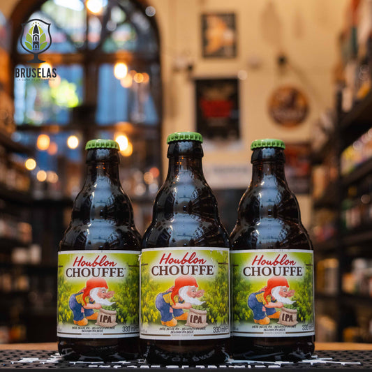 Botella de Houblon Chouffe, una Belgian IPA de Brasserie d'Achouffe. Con un ABV del 9%, esta cerveza combina el estilo belga con la intensidad del lúpulo. Presenta aromas cítricos, como toronja, y toques herbales y especiados gracias al uso de lúpulos Tomahawk, Saaz y Amarillo. Su sabor es amargo e intenso, equilibrado con una base maltosa y un final seco y refrescante. Ideal para maridar con bisque de langosta, hígados de pollo salteados o quesos azules.