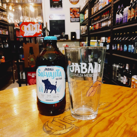 Vaso Jabalí 350ml, de cristal tipo pinta con logo serigrafiado en blanco, junto a una botella de Jabalí Salvajita Ultra. Ubicado sobre una barra en un ambiente cervecero, resaltando su uso ideal para cervezas artesanales mexicanas