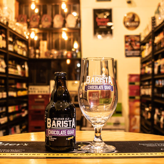 Copa Kasteel Barista Chocolate Quad 330ml, de cristal estilizado con logo blanco y morado, junto a una botella de Barista Chocolate Quad. Ubicada sobre una barra en un ambiente cervecero, resaltando su uso ideal para cervezas belgas oscuras.