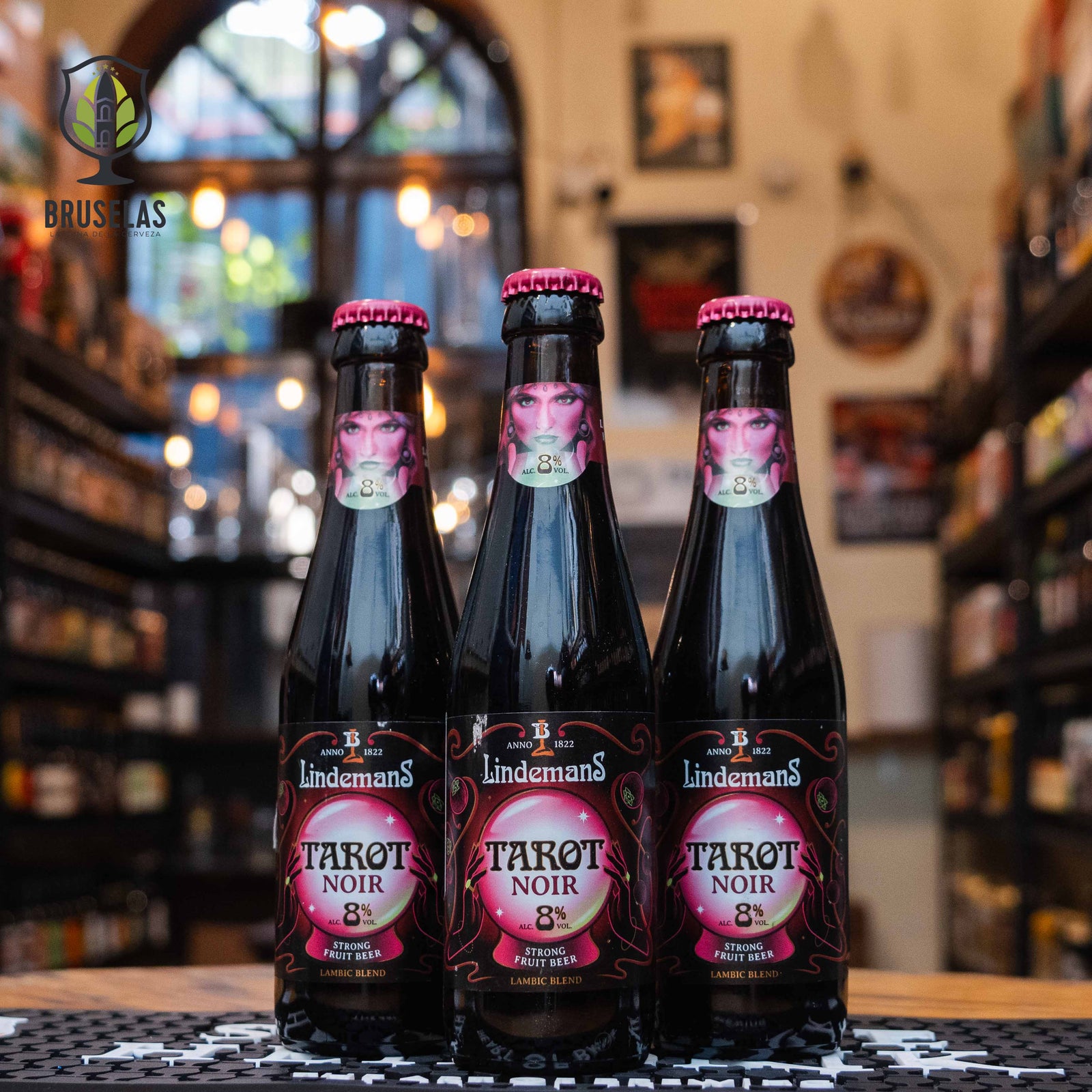 Botella de Tarot Noir de Lindemans, una Lambic Fruit belga con un ABV de 8%. La etiqueta presenta un diseño oscuro y místico, con la imagen de una mujer en tonos rosados y púrpura, evocando el arte del tarot. Esta cerveza, con sabor a bayas negras, equilibra dulzura y acidez. Ideal para maridar con carnes rojas o postres de chocolate. El diseño refleja su carácter misterioso y afrutado.