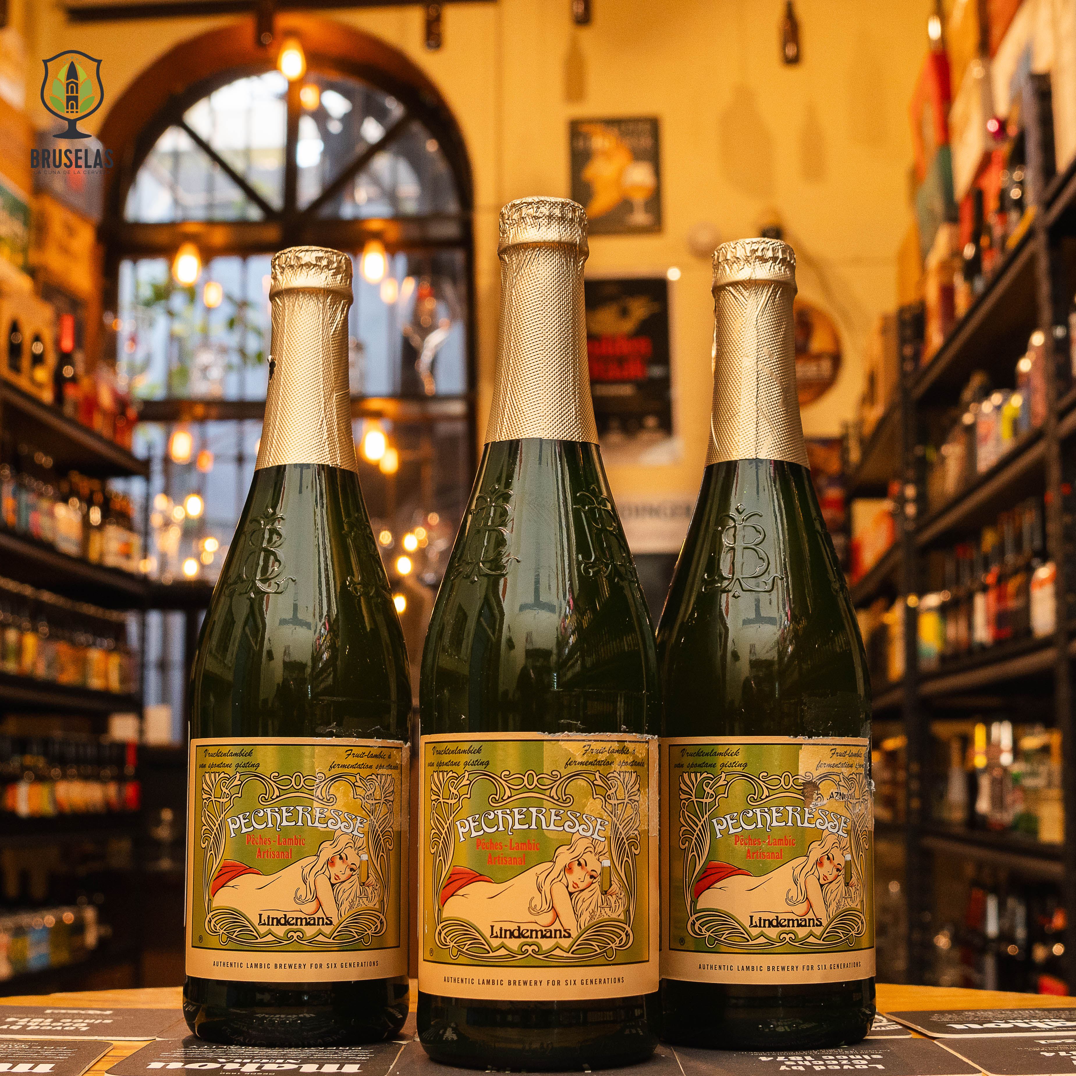 Botella de Lindemans Pecheresse, una Lambic Fruit belga de la cervecería Lindemans, ubicada en Pajottenland. Con un ABV de 2.5%, tiene un color dorado y burbujeo fino. Elaborada con jugo de durazno, ofrece aromas frutales con notas dominantes de durazno fresco. Su sabor es dulce, afrutado y ligeramente ácido. Ideal para maridar con ensaladas frescas, platos de cerdo, quesos suaves y postres de frutas.
