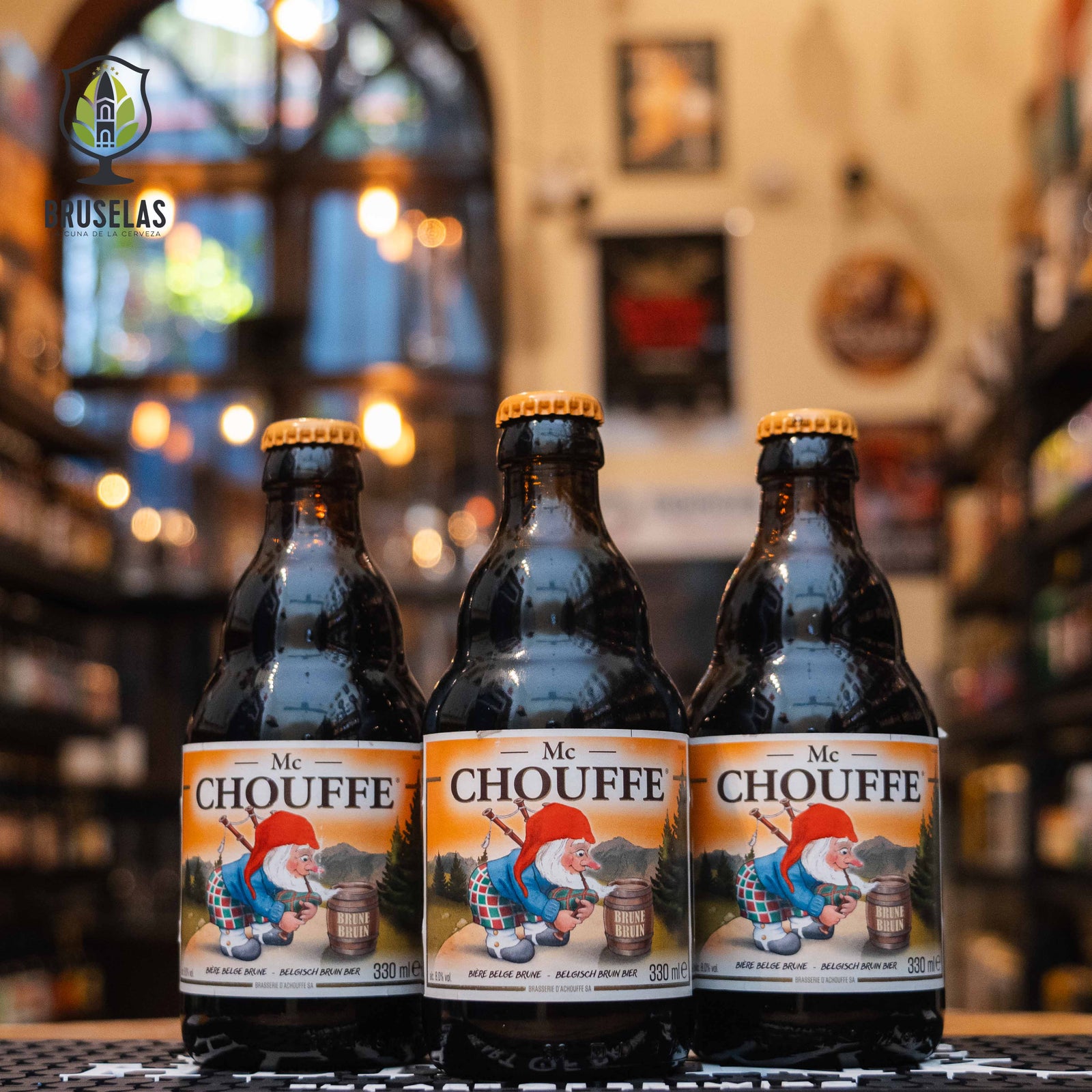 Botella de Mc Chouffe, una Belgian Strong Dark Ale de estilo Scotch Ale elaborada por Brasserie d'Achouffe. Con un ABV de 8%, esta cerveza oscura presenta un color rubí profundo y una espuma cremosa beige. Su aroma combina notas de caramelo, regaliz, anís y frutas oscuras como peras y ciruelas. En boca, ofrece un equilibrio entre dulzor de caramelo, especias y un final ligeramente amargo. Ideal para maridar con carnes asadas, jamón de Parma, quesos maduros y mariscos como salmón y ostras.