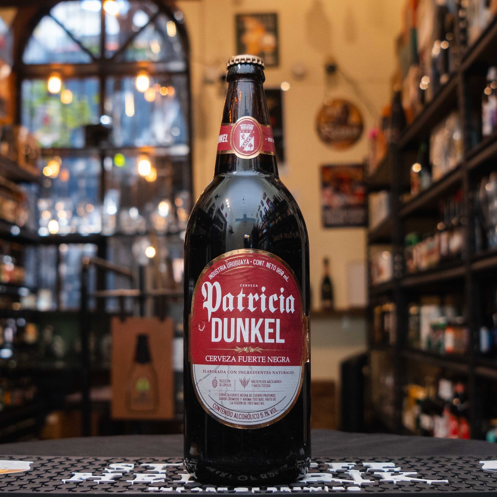 Botella de Patricia Dunkel, una cerveza negra estilo Dunkel de origen uruguayo. La etiqueta es roja con detalles blancos y negros, con letras clásicas que destacan el nombre. Con un ABV de 5.1%, ofrece notas de malta tostada, caramelo y un toque de chocolate. Ideal para maridar con carnes a la parrilla, embutidos y quesos maduros. Su diseño y formato caguama la hacen perfecta para compartir momentos especiales.