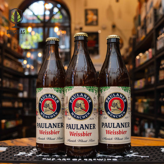 Botella de Paulaner Weissbier, una cerveza de trigo estilo Weissbier elaborada por Paulaner Brauerei, Múnich, Alemania. La etiqueta tiene el icónico logo de un monje en un círculo rojo y azul. Presenta un ABV de 5.5%, con sabores suaves y frutales, destacando plátano y clavo. Ideal para acompañar ensaladas, pescados y salchichas bávaras.