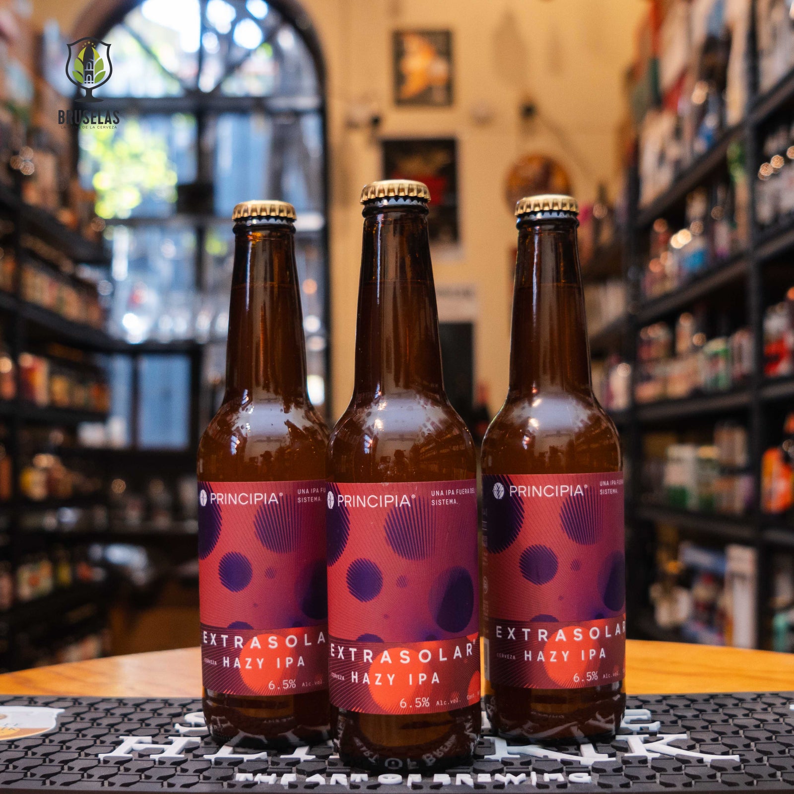 Principia Extrasolar Hazy IPA, una Hazy IPA de Principia. Su etiqueta presenta un diseño con tonos morados y rojos, simulando un paisaje cósmico con planetas y nebulosas. Su perfil aromático destaca notas de mango, piña y durazno, con un cuerpo sedoso y jugoso. Ideal para maridar con tacos de pescado, sushi, alitas picantes y quesos suaves.