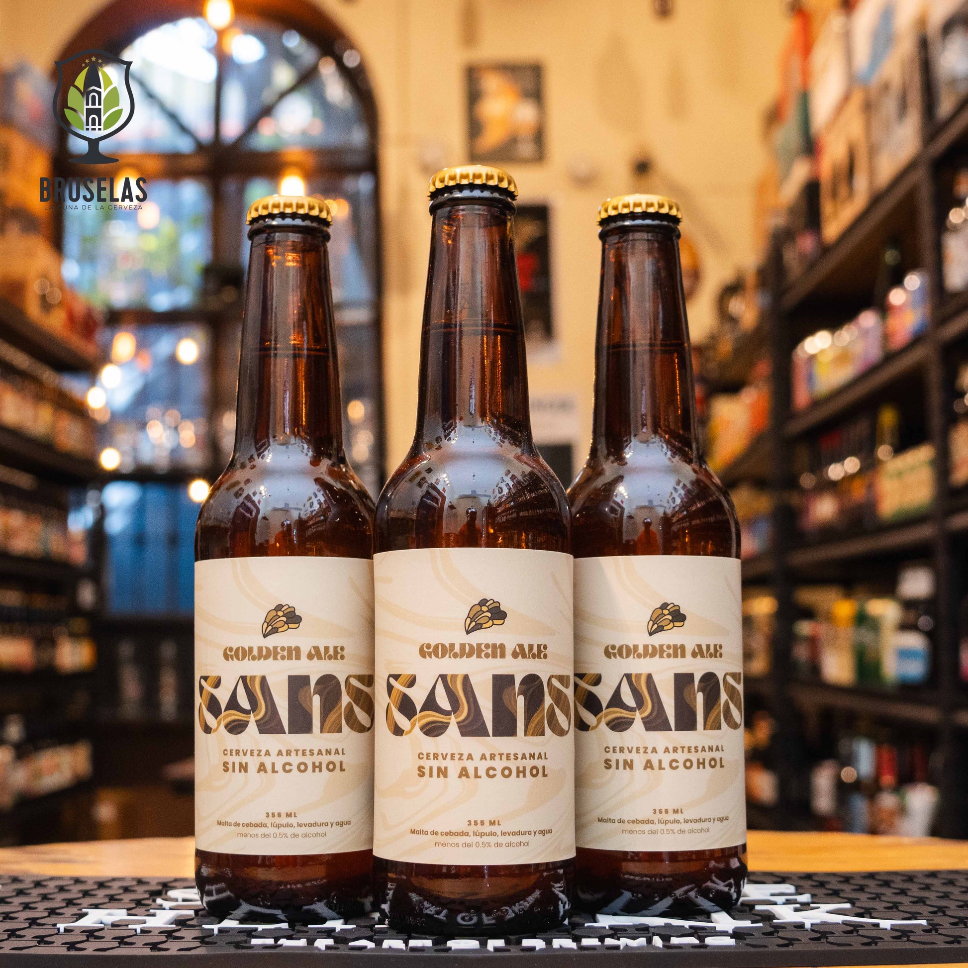 Botella de Sans Golden Ale Sin Alcohol, una cerveza artesanal estilo Golden Ale de la Cervecería SANS, México. La etiqueta es beige con detalles dorados. ABV de 0.0%, con aromas de maltas suaves y notas cítricas. Sabor ligero y equilibrado con un final refrescante. Ideal para acompañar ensaladas, pescados y platos ligeros. Diseño elegante y refrescante.