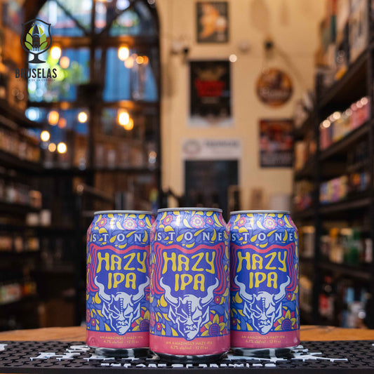 Lata de Stone Hazy IPA, una Hazy IPA de Stone Brewing, elaborada en EE.UU. La etiqueta es colorida con tonos púrpura y amarillo, decorada con flores y un logo de gárgola en blanco. Con un ABV de 6.7%, ofrece sabores tropicales de mango, piña, melón y un toque cítrico. Ideal para maridar con pollo glaseado, ceviche con mango y ensaladas frutales. El diseño de la lata es vibrante y moderno, evocando frescura y diversión.