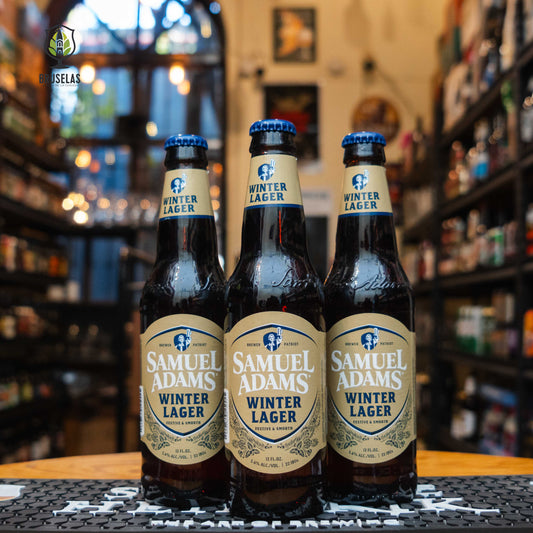 Botellas de Samuel Adams Winter Lager, una Bock invernal con 5.6% ABV, con etiqueta en tonos beige y azul. Destaca por sus notas de caramelo, naranja y especias, ideal para maridar con carnes asadas y postres de temporada.