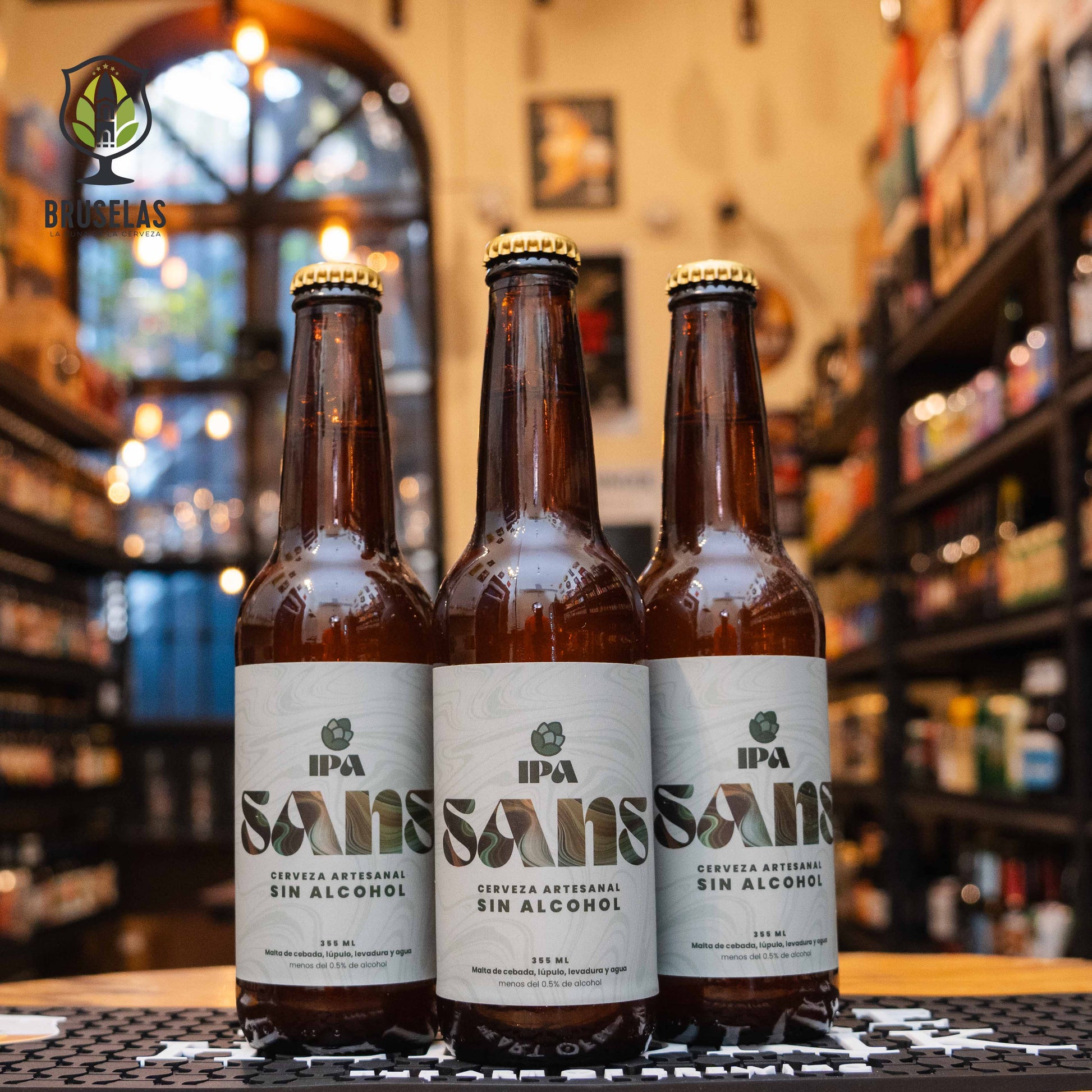 Botella de Sans IPA Sin Alcohol, una cerveza artesanal estilo IPA de la Cervecería SANS, México. La etiqueta es blanca con detalles modernos. ABV de 0.0%, con aromas frutales de mango y cítricos. Sabor tropical con notas de mango y cítricos, y un final sutilmente amargo. Ideal para acompañar carnes a la parrilla, mariscos y platos especiados. Diseño innovador y refrescante.