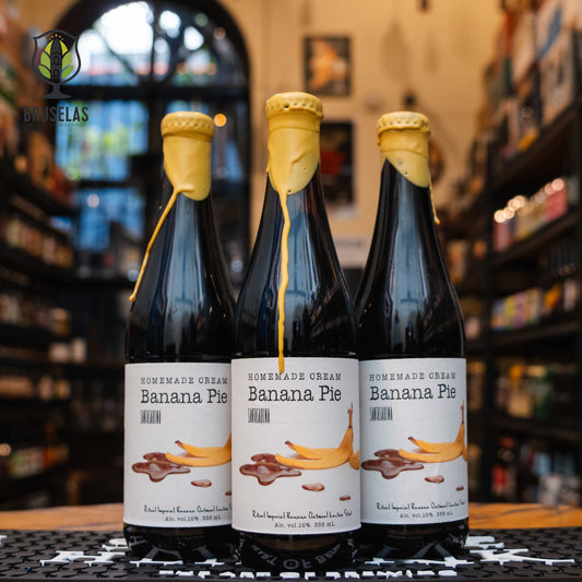 Botella de Santa Sabina Homemade Cream Banana Pie, una Pastry Ale de Santa Sabina Brewery. La etiqueta es blanca con un diseño de plátano y chocolate derretido. ABV de 10%, con aromas intensos de plátano maduro y vainilla. Sabor dulce y cremoso con notas de plátano, vainilla y caramelo. Ideal para acompañar cheesecake, pasteles de frutas y helados. Diseño atractivo y postrero.