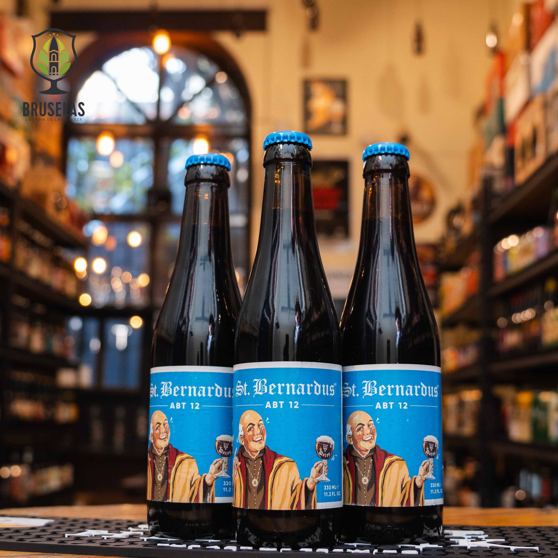 Botella de St. Bernardus Abt 12, una Quadrupel de la cervecería Brouwerij St. Bernardus, con un ABV de 10%. La etiqueta presenta un monje sonriente con fondo azul claro, sosteniendo una copa de cerveza. De color marrón oscuro con reflejos rubí, ofrece aromas de frutas oscuras como ciruelas y pasas, acompañados de caramelo y especias. Su sabor es complejo y suave, ideal para maridar con carnes asadas y quesos fuertes.