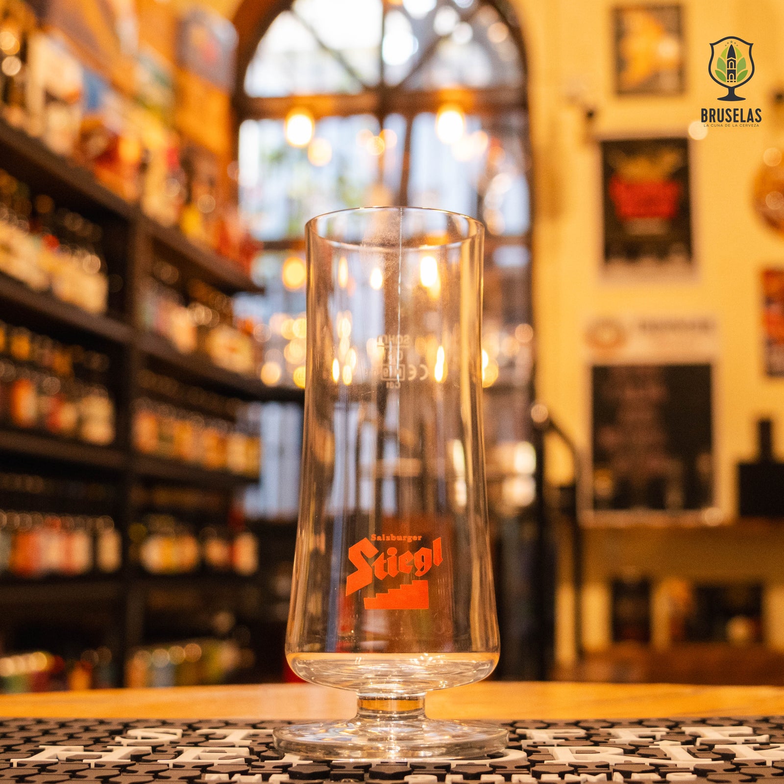 Vaso Stiegl 300ml, de cristal alto y acampanado con logo rojo, junto a una lata de Stiegl Radler Zitrone. Ubicado sobre una barra en un ambiente cervecero, resaltando su uso ideal para cervezas lager y radler austríacas.