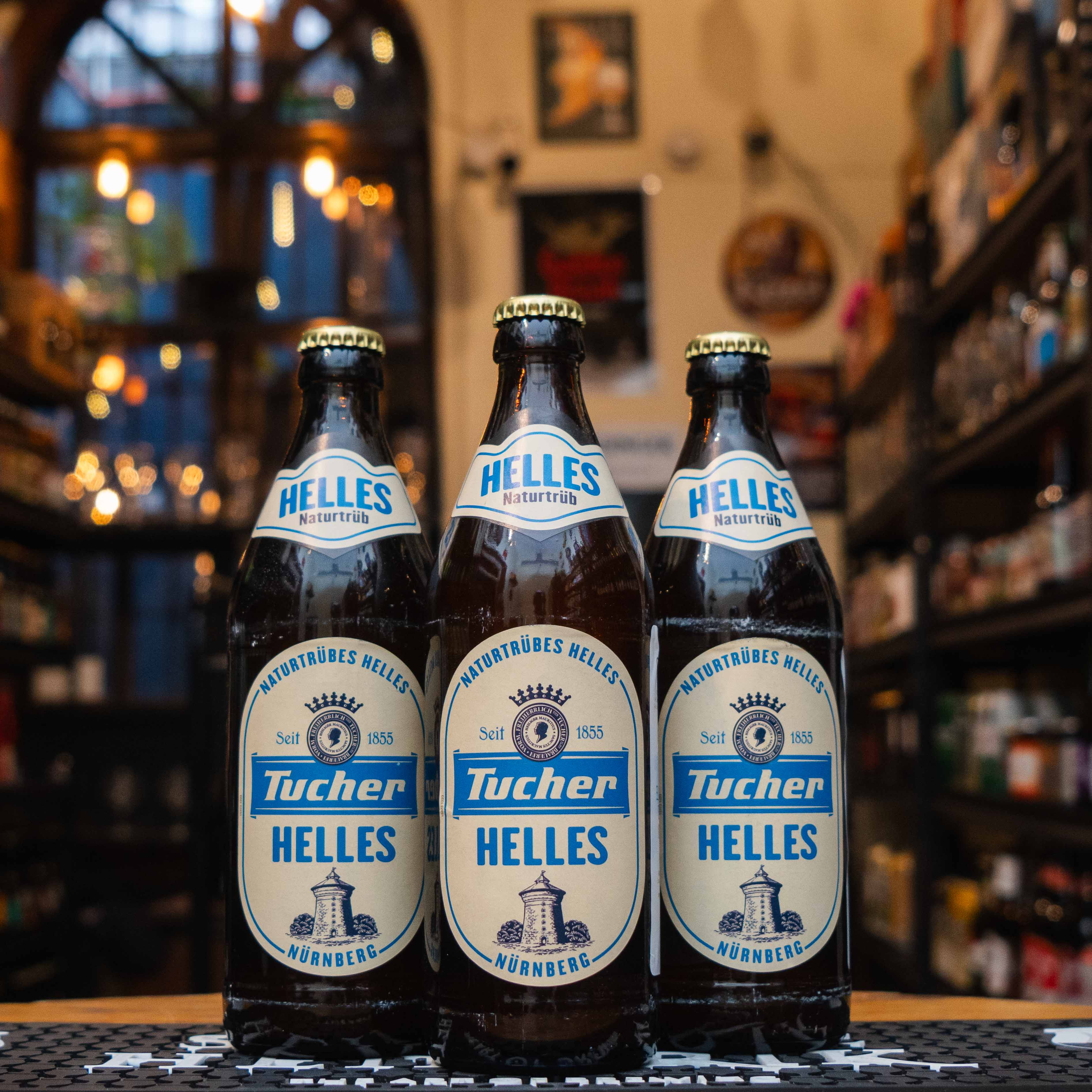 Botella de Tucher Helles Naturtrüb, una lager alemana sin filtrar con 5.2% ABV, elaborada por Tucher Bräu en Núremberg. Su etiqueta destaca tonos beige y azul, con el logotipo de la cervecería y detalles clásicos. Ofrece notas de pan fresco, cereales y un toque herbal, con un final maltoso y refrescante. Ideal para maridar con salchichas bávaras, pretzels y quesos suaves. Tradición y autenticidad en cada sorbo.