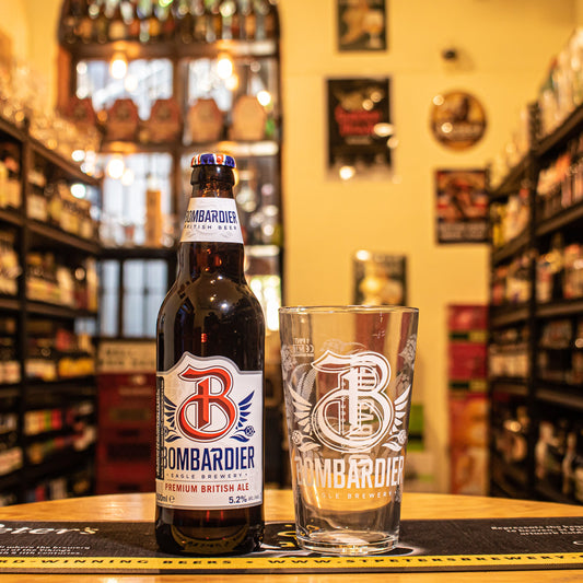 Vaso Bombardier 500ml, de cristal tipo pinta con logo serigrafiado en blanco, junto a una botella de Bombardier Premium British Ale. Capturado en una barra con fondo cervecero, resaltando su uso ideal para degustar ales británicas.