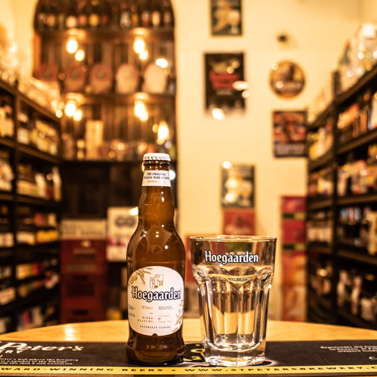 Vaso Hoegaarden 250ml, de cristal hexagonal con logo en blanco, junto a una botella de Hoegaarden Witbier. Ubicado sobre una barra en un ambiente cervecero, resaltando su uso ideal para cervezas de trigo belgas.