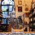 Vaso Mikkeller 400ml, de cristal transparente con el icónico logo negro de la marca. Ubicado sobre una barra con posavasos de goma, en un ambiente cervecero con estanterías llenas de botellas en el fondo, reflejando su uso en bares o cervecerías.
