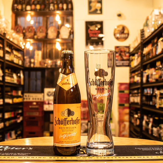 Vaso Schöfferhofer Weizen 500ml, de cristal alto y estilizado con logo negro y dorado, junto a una botella de Schöfferhofer Hefeweizen. Ubicado sobre una barra en un ambiente cervecero, resaltando su uso ideal para cervezas de trigo alemanas.