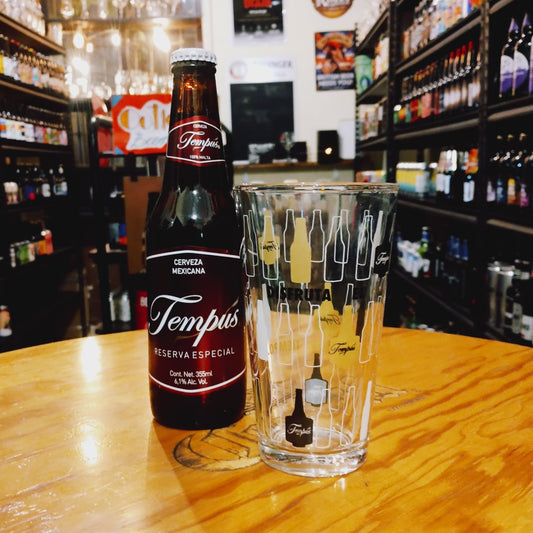 Vaso Tempus Chelero 350ml, de cristal tipo pinta con ilustraciones de botellas y logo Tempus, junto a una botella de Tempus Reserva Especial. Ubicado sobre una barra en un ambiente cervecero, resaltando su uso ideal para cervezas artesanales mexicanas.