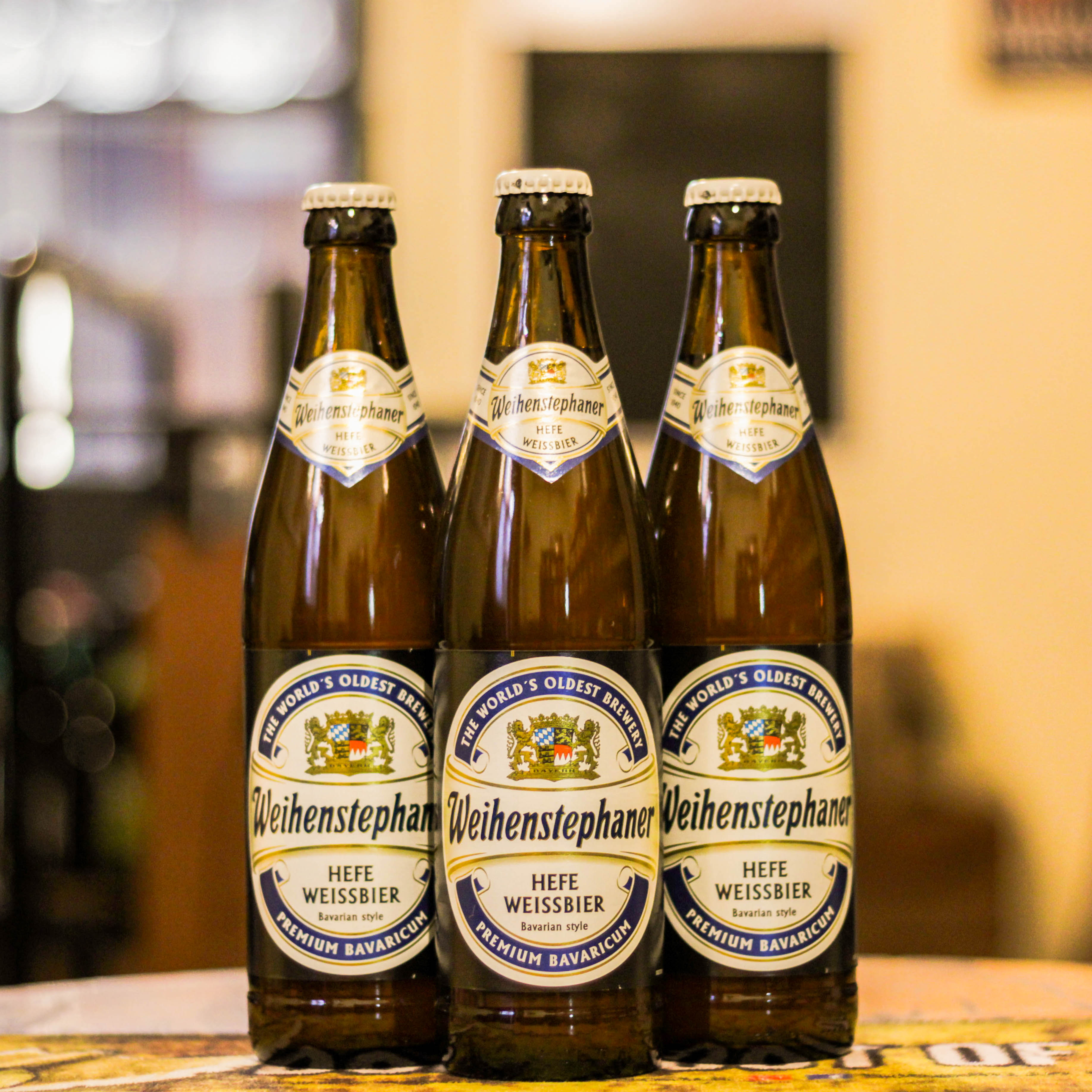 Botella de Weihenstephaner Hefeweissbier, una Hefeweizen de la cervecería Bayerische Staatsbrauerei Weihenstephan con un ABV de 5.4%. Ofrece aromas a plátano, clavo y levadura, con un sabor suave y cremoso, equilibrado entre notas frutales y especiadas. Ideal para maridar con salchichas bávaras, quesos especiados y mariscos, destacando su refrescante y ligera textura.