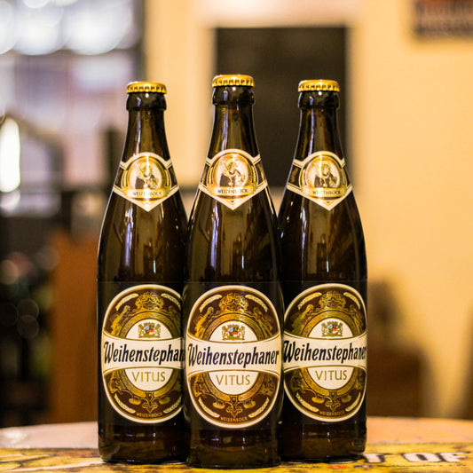 Botella de Weihenstephaner Vitus, una Weizenbock elaborada por la cervecería Weihenstephan en Alemania. La etiqueta presenta el escudo dorado con detalles en azul y blanco. ABV de 7.7%, con sabores afrutados de plátano y albaricoque, y notas de clavo. Ideal para acompañar postres de frutas. Refleja la tradición de la cervecería más antigua del mundo, perfecta para cualquier época del año.