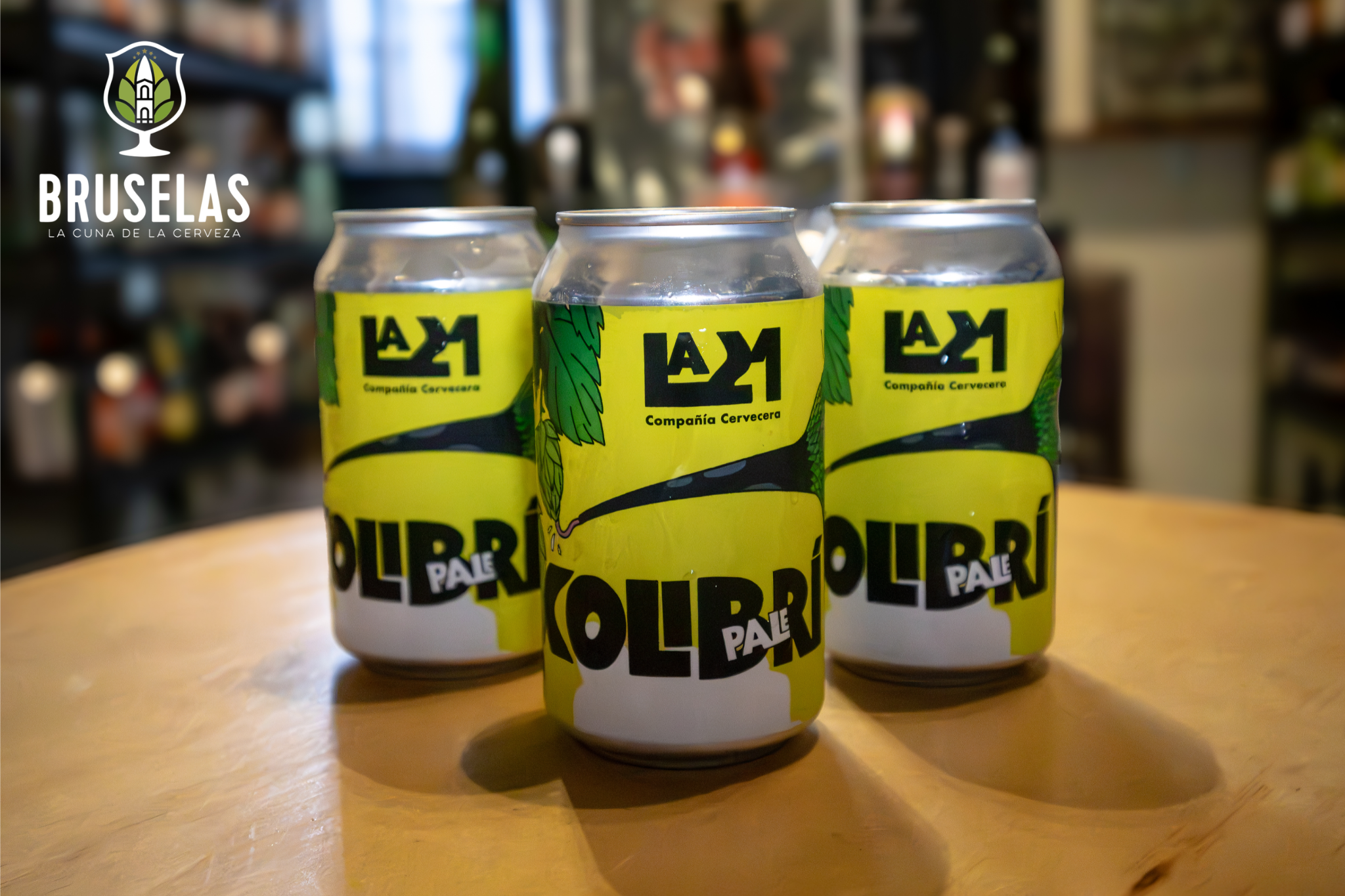 tres latas de cerveza artesanal La 21 que lleva por nombre La Kolibri, sobre una mesa de madera con el fondo desenfocado en una beer boutique