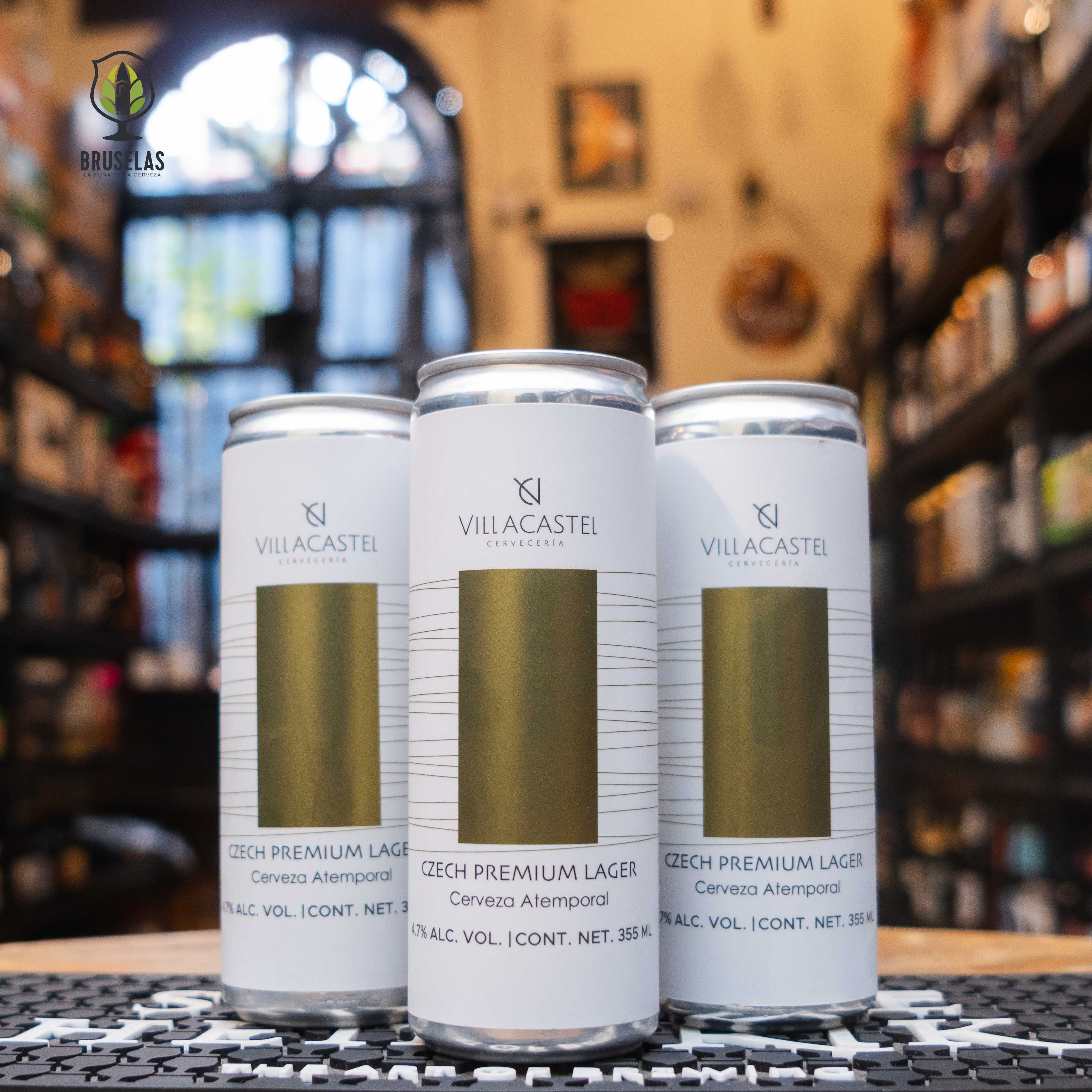 “Czech Premium Lager” de Villacastel en un diseño minimalista y sofisticado, con una franja dorada al centro y fondo blanco elegante. Las latas están sobre una barra con fondo desenfocado de tienda especializada, lo que transmite un ambiente exclusivo y artesanal.