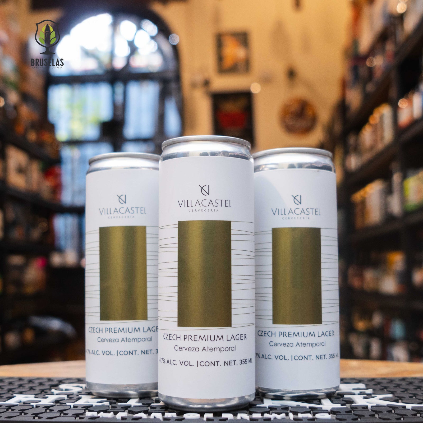 “Czech Premium Lager” de Villacastel en un diseño minimalista y sofisticado, con una franja dorada al centro y fondo blanco elegante. Las latas están sobre una barra con fondo desenfocado de tienda especializada, lo que transmite un ambiente exclusivo y artesanal.