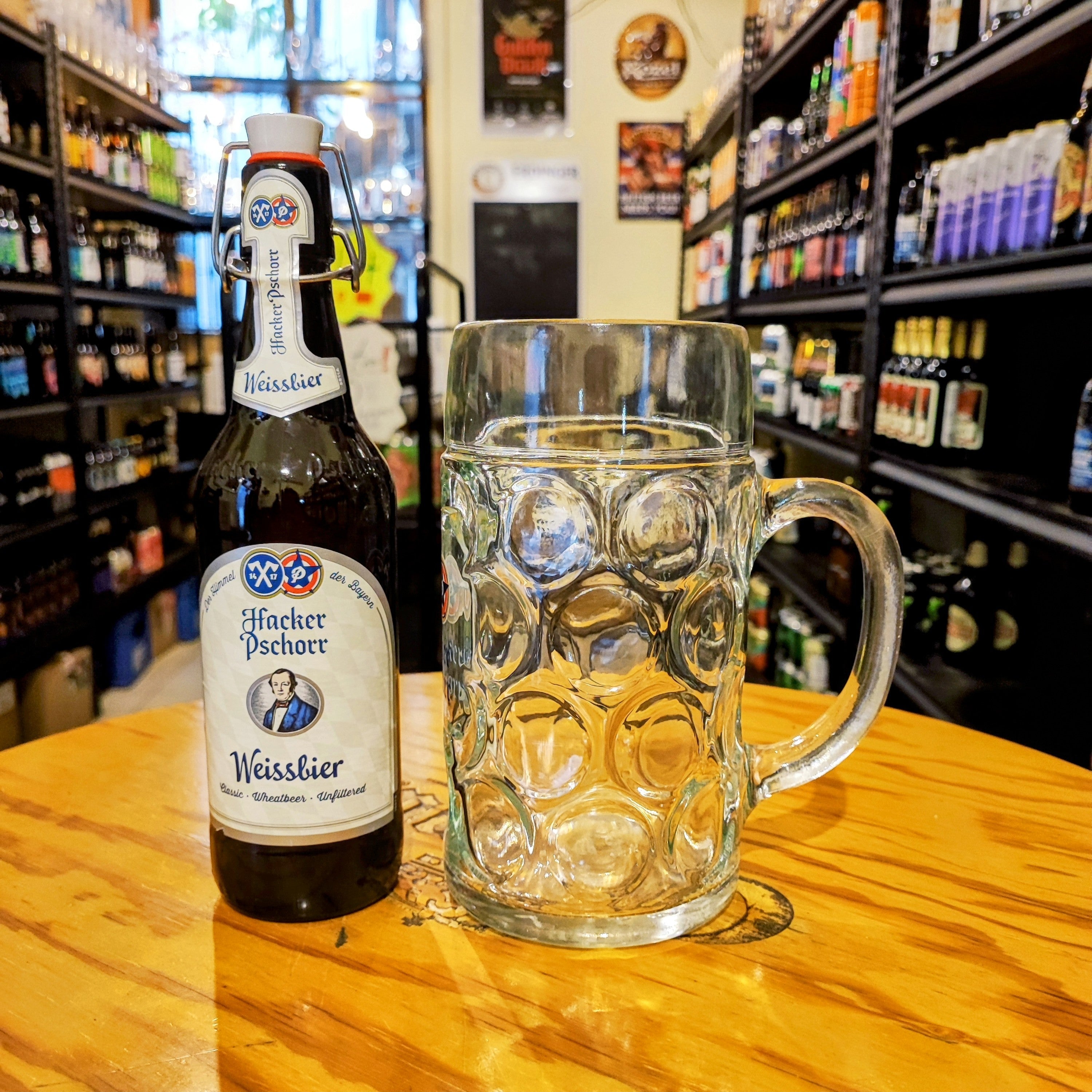 Tarro Hacker Pschorr de 1 litro junto a una botella de Weissbier, sobre mesa en tienda de cervezas artesanales