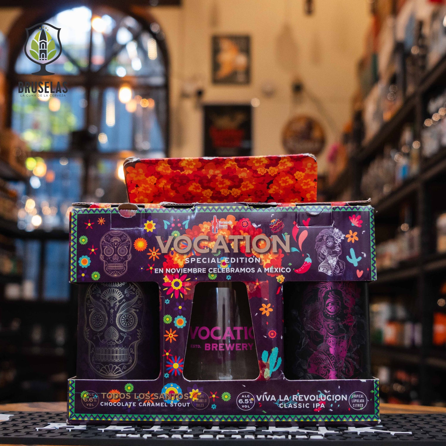 El Giftpack Vocation "Viva la Revolución" destaca con su vibrante diseño inspirado en la cultura mexicana, con calaveras y motivos florales en tonos morados y naranjas. Contiene dos cervezas en latas decoradas y un vaso con el logo de Vocation en color morado. La caja tiene una ventana que permite ver los productos, y el fondo muestra una tienda de cervezas artesanales con iluminación cálida. Cambios sujetos a disponibilidad.